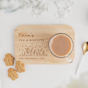 K&ouml;nnte beinhalten: Ein Holz-Teetablett mit den Worten "Nana's TEA & BISCUITS" eingraviert. Das Tablett enth&auml;lt eine transparente Glastasse Tee, einen goldenen L&ouml;ffel und drei Lebkuchenkekse. Das Tablett hat auch ein Blumendesign und die Worte "LOVE FROM ROSIE & THEO".
