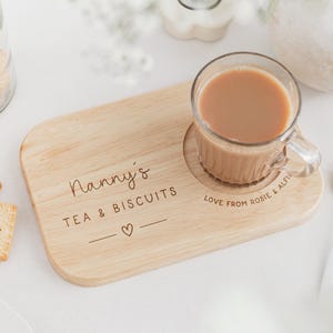 May include: A wooden tea and biscuit serving board with the text "Nanny's TEA & BISCUITS" and a heart symbol. A glass mug of tea sits on the board, next to a gold spoon. Biscuits are in a glass jar.