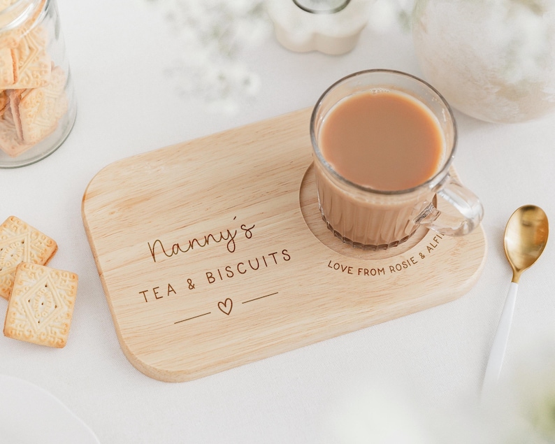 May include: A wooden tea and biscuit serving board with the text "Nanny's TEA & BISCUITS" and a heart symbol. A glass mug of tea sits on the board, next to a gold spoon. Biscuits are in a glass jar.