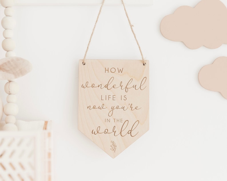 May include: Wooden sign with the text "How wonderful life is now you're in the world" in a cursive font. The sign is hanging from a string and has a small floral design at the bottom.