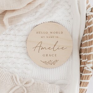 May include: A wooden round sign with the text "Hello World, My Name Is Amelie Grace" and a small floral design.