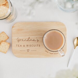 May include: A wooden serving board with the text "Grandma's TEA & BISCUITS" and a heart symbol. A clear glass mug with tea sits on the board. A gold and white spoon is next to the board, and square biscuits are nearby.