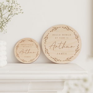 May include: Two round wooden signs with the text "HELLO WORLD MY NAME IS Arthur JAMES". The signs are light brown with a floral wreath design around the edge. One sign is smaller than the other.