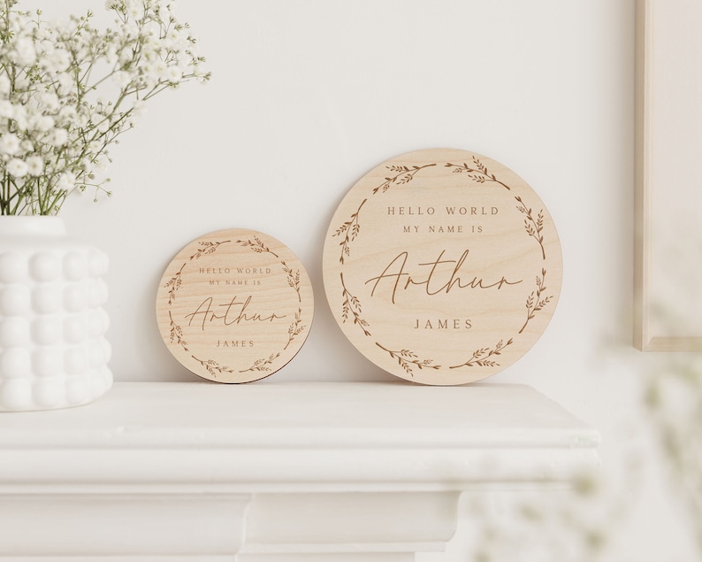 May include: Two round wooden signs with the text "HELLO WORLD MY NAME IS Arthur JAMES". The signs are light brown with a floral wreath design around the edge. One sign is smaller than the other.