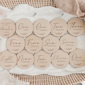May include: A set of 12 wooden baby milestone discs with engraved text, including "Hello World, My Name Is Amelie Grace", "One Week", "One Month", "Two Months", "Three Months", "Four Months", "Five Months", "Six Months", "Seven Months", "Eight Months", "Nine Months", "Ten Months", "Eleven Months", and "One Year Old".