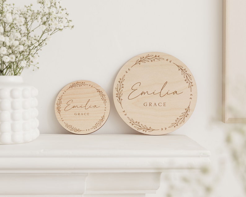 May include: Two round wooden signs with a floral design and the name "Emilia Grace" engraved on them. The signs are different sizes and are displayed on a white shelf.