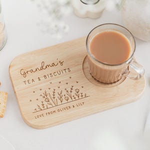 May include: A wooden tea and biscuit serving board with a clear glass mug of tea. The board is engraved with "Grandma's TEA & BISCUITS" and "LOVE FROM OLIVER & LILY". A gold spoon and biscuits are also present.