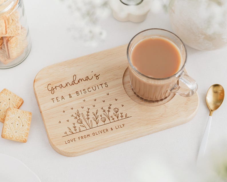 May include: A wooden tea and biscuit serving board with a clear glass mug of tea. The board is engraved with "Grandma's TEA & BISCUITS" and "LOVE FROM OLIVER & LILY". A gold spoon and biscuits are also present.