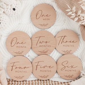 May include: Set of six wooden baby milestone discs with a natural wood finish. Each disc features a different age milestone, from "One Week" to "Six Months", with a simple floral design.