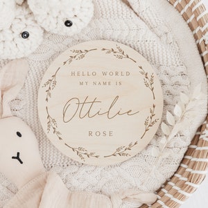 May include: A round wooden sign with the text "HELLO WORLD MY NAME IS Ottilie ROSE" in a decorative font, surrounded by a floral wreath. The sign is on a white knitted blanket, with a stuffed bunny and baby booties in the background.