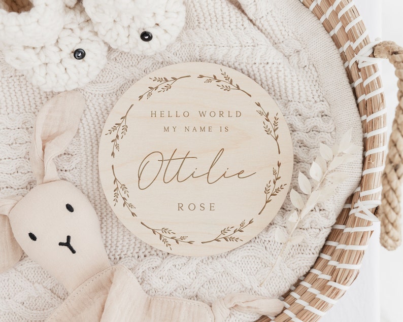 May include: A round wooden sign with the text "HELLO WORLD MY NAME IS Ottilie ROSE" in a decorative font, surrounded by a floral wreath. The sign is on a white knitted blanket, with a stuffed bunny and baby booties in the background.