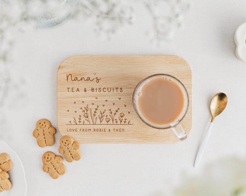 May include: A wooden tea and biscuit tray with the text "Nana's TEA & BISCUITS" and floral engraving. A clear glass mug with tea, three gingerbread cookies, and a gold and white spoon are on a white surface.