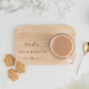 May include: A wooden serving tray with the text "Dad's TEA & BISCUITS" and a star. A clear glass mug of tea sits on the tray, next to a gold spoon and gingerbread cookies. The text "Love from Rach" is also visible.