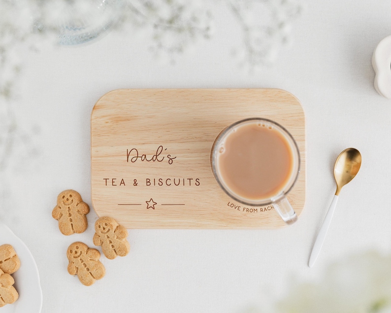 May include: A wooden serving tray with the text "Dad's TEA & BISCUITS" and a star. A clear glass mug of tea sits on the tray, next to a gold spoon and gingerbread cookies. The text "Love from Rach" is also visible.
