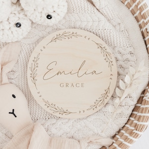 May include: A round wooden plaque with a floral design and the name "Emilia Grace" engraved in cursive script.