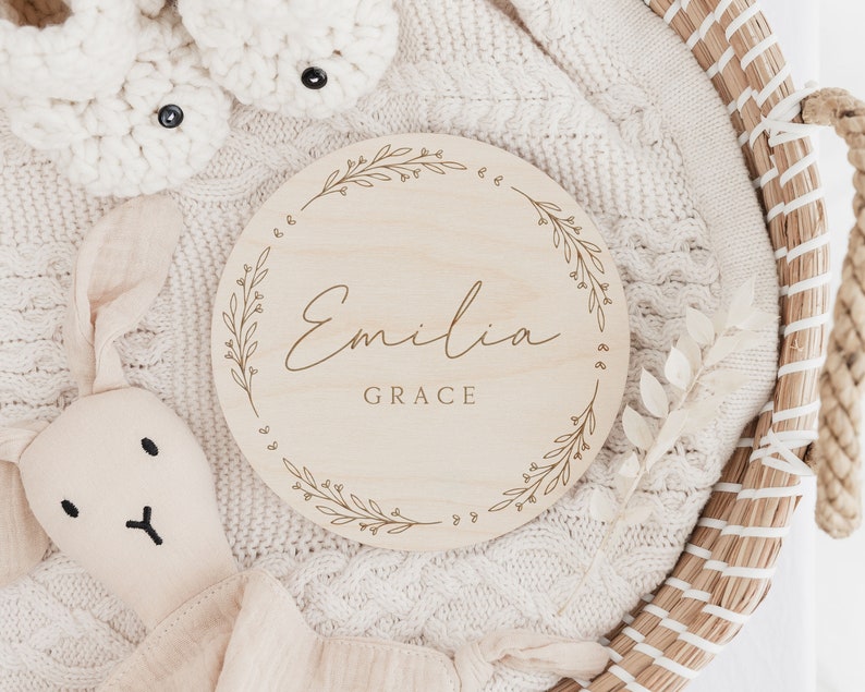 May include: A round wooden plaque with a floral design and the name "Emilia Grace" engraved in cursive script.