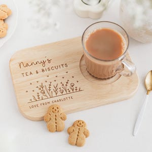 May include: A wooden tea and biscuit serving board with the text "Nanny's TEA & BISCUITS" and "LOVE FROM CHARLOTTE". A clear glass mug with tea, two gingerbread men cookies, and a gold and white spoon are on the white surface.