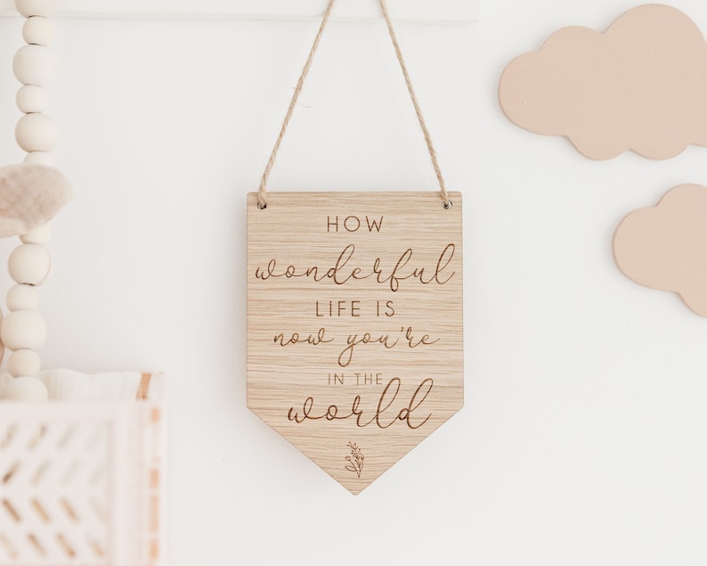 May include: Wooden sign with the text "How wonderful life is now you're in the world" in a cursive font. The sign is hanging from a string and has a small floral design at the bottom.