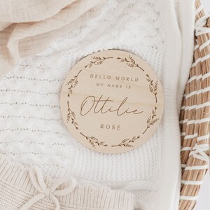 May include: A round wooden baby announcement sign with the text "HELLO WORLD MY NAME IS Ottilie ROSE" in a decorative font. The sign is surrounded by a floral wreath design. The sign is resting on a white knitted blanket.