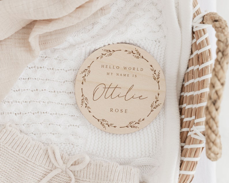 May include: A round wooden baby announcement sign with the text "HELLO WORLD MY NAME IS Ottilie ROSE" in a decorative font. The sign is surrounded by a floral wreath design. The sign is resting on a white knitted blanket.