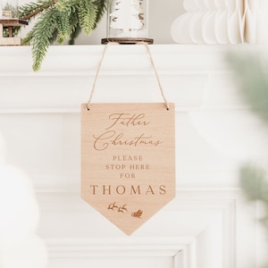 Op de afbeelding: Een houten bord met de tekst "Kerstman Stop hier alstublieft voor THOMAS" en een klein beeld van een slee met rendieren. Het bord hangt aan een schoorsteenmantel.