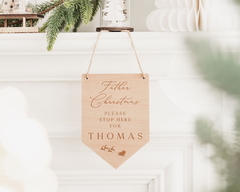 Op de afbeelding: Een houten bord met de tekst "Kerstman Stop hier alstublieft voor THOMAS" en een klein beeld van een slee met rendieren. Het bord hangt aan een schoorsteenmantel.