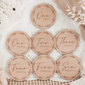 May include: A set of seven wooden milestone discs for baby photos. Each disc is round with a decorative leaf border and features text indicating time increments from one week to six months.