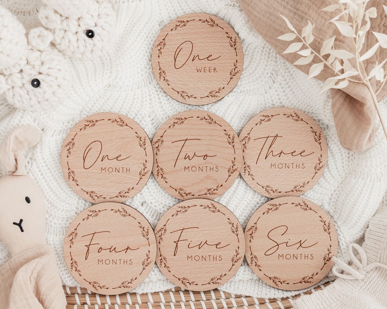 May include: A set of seven wooden milestone discs for baby photos. Each disc is round with a decorative leaf border and features text indicating time increments from one week to six months.