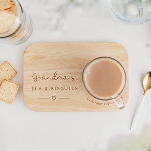 K&ouml;nnte beinhalten: Ein Holzservierbrett mit dem Text "Grandma's TEA & BISCUITS" und einem Herzsymbol. Eine klare Glastasse mit Tee steht auf dem Brett. Ein goldener und wei&szlig;er L&ouml;ffel liegt neben dem Brett, und quadratische Kekse sind in der N&auml;he.