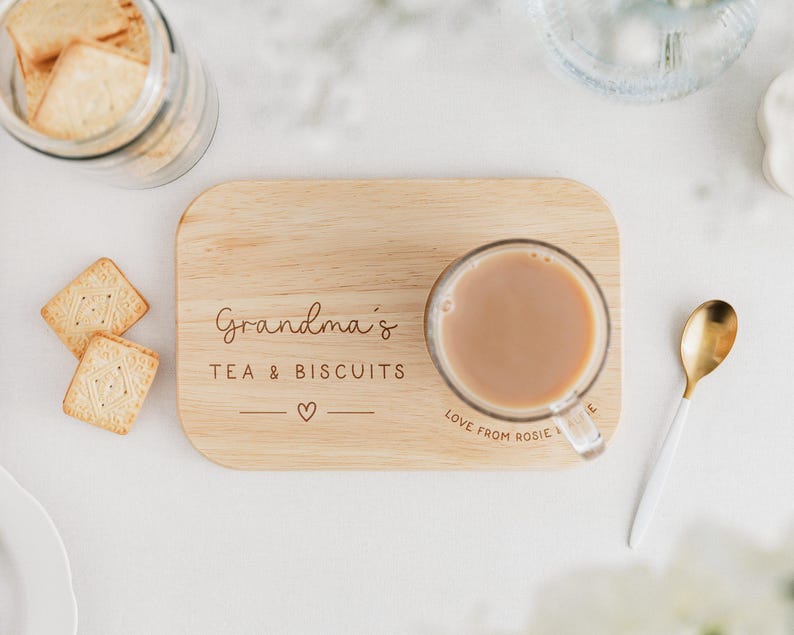 K&ouml;nnte beinhalten: Ein Holzservierbrett mit dem Text "Grandma's TEA & BISCUITS" und einem Herzsymbol. Eine klare Glastasse mit Tee steht auf dem Brett. Ein goldener und wei&szlig;er L&ouml;ffel liegt neben dem Brett, und quadratische Kekse sind in der N&auml;he.