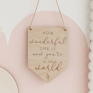 May include: Wooden sign with the text "How wonderful life is now you're in the world" in a cursive font. The sign is hanging from a string and has a small floral design at the bottom.
