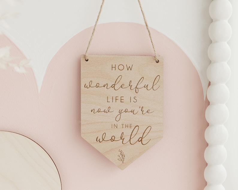 May include: Wooden sign with the text "How wonderful life is now you're in the world" in a cursive font. The sign is hanging from a string and has a small floral design at the bottom.