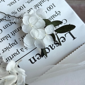 Puede incluir: Una horquilla con delicados p&eacute;talos de hortensia blancos preservados y peque&ntilde;as hojas de color verde oscuro unidos a un tallo de alambre met&aacute;lico fino. El arreglo floral se sit&uacute;a sobre un papel blanco con texto impreso en negro.