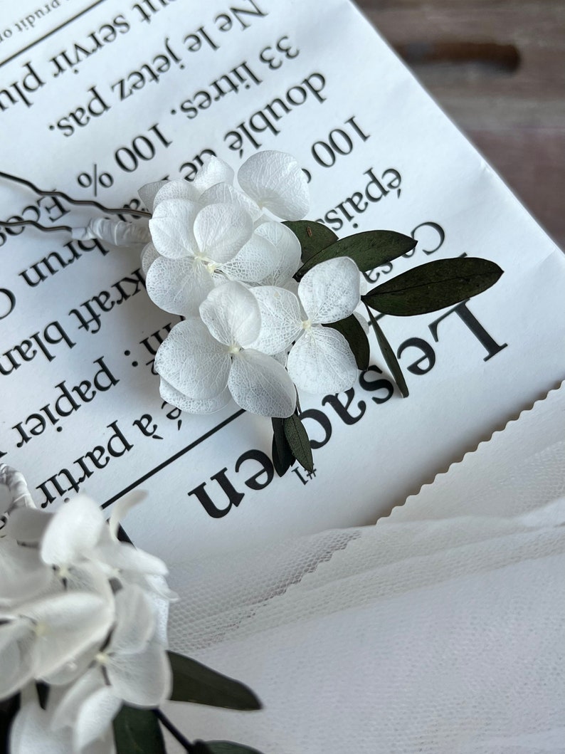 Puede incluir: Una horquilla con delicados p&eacute;talos de hortensia blancos preservados y peque&ntilde;as hojas de color verde oscuro unidos a un tallo de alambre met&aacute;lico fino. El arreglo floral se sit&uacute;a sobre un papel blanco con texto impreso en negro.