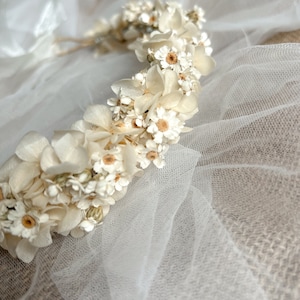May include: A floral crown featuring an arrangement of small white flowers and cream-colored hydrangea petals. The headpiece is constructed on a thin, flexible base and is accented with delicate white tulle fabric draped around the floral elements.