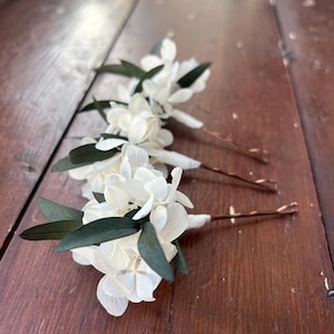 Puede incluir: Tres horquillas para el cabello con racimos de p&eacute;talos de hortensia blancos y hojas de color verde oscuro unidas a horquillas de metal color cobre. Cada arreglo floral est&aacute; asegurado con cinta floral blanca en la base del pasador.