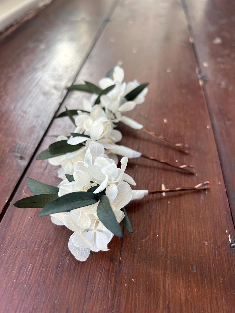 Puede incluir: Tres horquillas para el cabello con racimos de p&eacute;talos de hortensia blancos y hojas de color verde oscuro unidas a horquillas de metal color cobre. Cada arreglo floral est&aacute; asegurado con cinta floral blanca en la base del pasador.