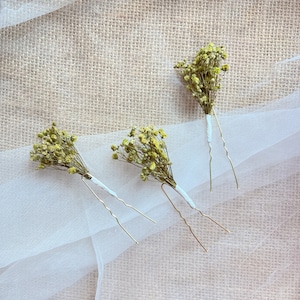 Op de afbeelding: Drie gouden haarspelden met gedroogde groene bloemen eraan. De spelden liggen op een witte stoffen achtergrond.