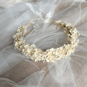 May include: A floral crown featuring cream and off-white dried hydrangea petals and small white blossoms with yellow centers. The circular arrangement is secured with a thin, light-colored cord and includes a sheer white ribbon tie at the back.