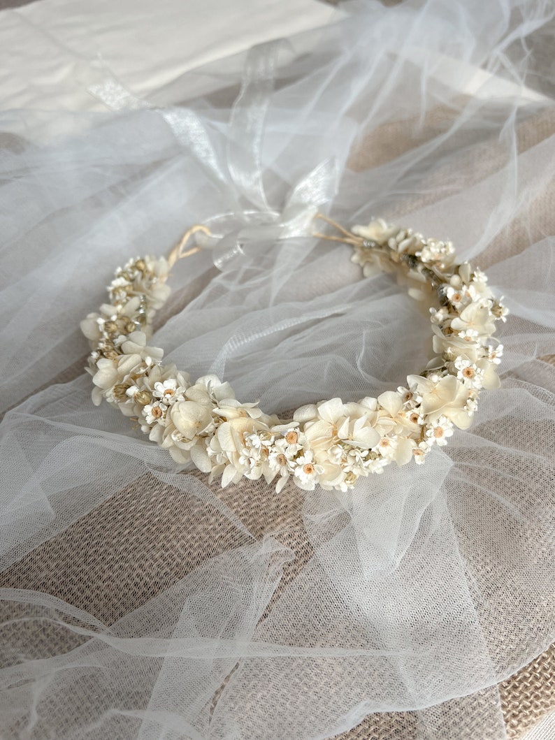 May include: A floral crown featuring cream and off-white dried hydrangea petals and small white blossoms with yellow centers. The circular arrangement is secured with a thin, light-colored cord and includes a sheer white ribbon tie at the back.