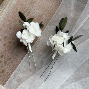 Puede incluir: Dos horquillas para el cabello con racimos de p&eacute;talos de hortensia blancos y hojas de color verde oscuro. Cada arreglo floral est&aacute; sujeto a una horquilla de metal plateado en forma de U con la base del tallo envuelta en blanco.
