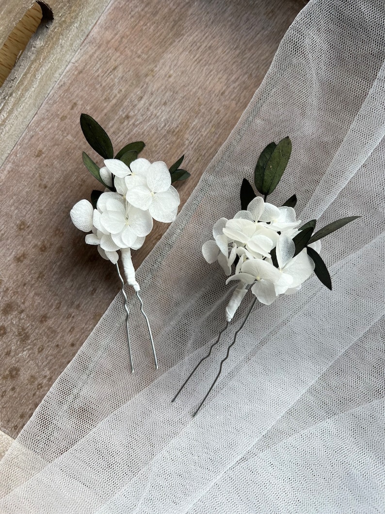 Puede incluir: Dos horquillas para el cabello con racimos de p&eacute;talos de hortensia blancos y hojas de color verde oscuro. Cada arreglo floral est&aacute; sujeto a una horquilla de metal plateado en forma de U con la base del tallo envuelta en blanco.