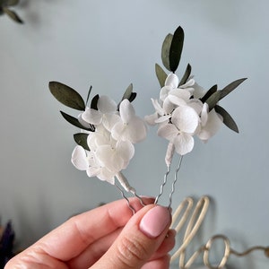 Puede incluir: Dos horquillas para el cabello con racimos de peque&ntilde;os p&eacute;talos de hortensia blancos y hojas de color verde oscuro. Cada arreglo floral est&aacute; sujeto a una horquilla de metal ondulado plateado, con los tallos envueltos en cinta floral blanca.