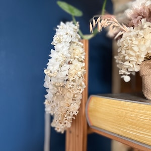 May include: A circular floral crown composed of dried white and cream-colored hydrangea blossoms and small filler flowers. The delicate arrangement features layered petals in soft, neutral tones, creating a dense, textured wreath structure.