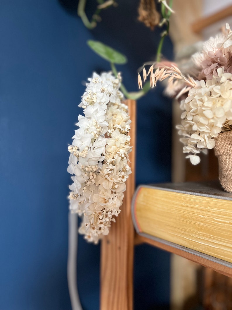 May include: A circular floral crown composed of dried white and cream-colored hydrangea blossoms and small filler flowers. The delicate arrangement features layered petals in soft, neutral tones, creating a dense, textured wreath structure.