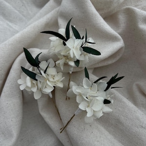 Puede incluir: Tres horquillas para el cabello con racimos de p&eacute;talos de hortensia blancos acentuados por peque&ntilde;as hojas de color verde oscuro. Cada arreglo floral est&aacute; sujeto a una fina horquilla de metal dorado, creando un delicado conjunto de accesorios.