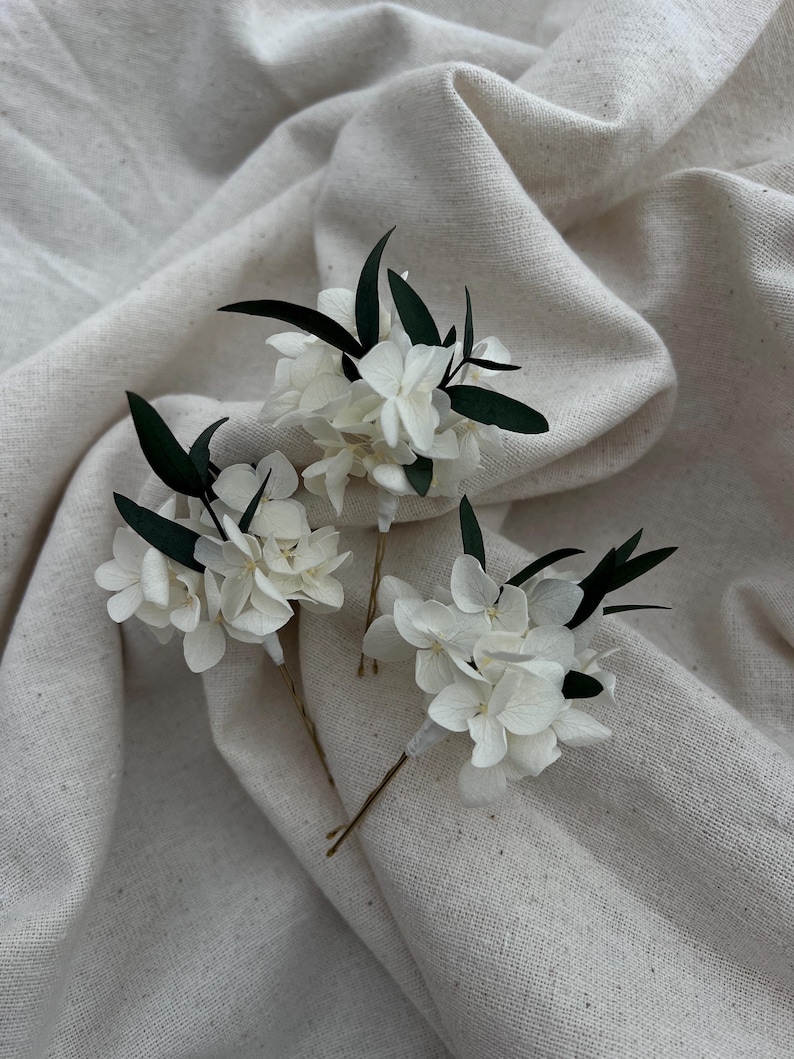 Puede incluir: Tres horquillas para el cabello con racimos de p&eacute;talos de hortensia blancos acentuados por peque&ntilde;as hojas de color verde oscuro. Cada arreglo floral est&aacute; sujeto a una fina horquilla de metal dorado, creando un delicado conjunto de accesorios.