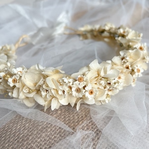 May include: A floral crown featuring an arrangement of cream and off-white dried flowers, including small daisies and hydrangea-like petals. The circular headpiece is constructed with a thin, flexible base and includes white ribbon ties at the back.