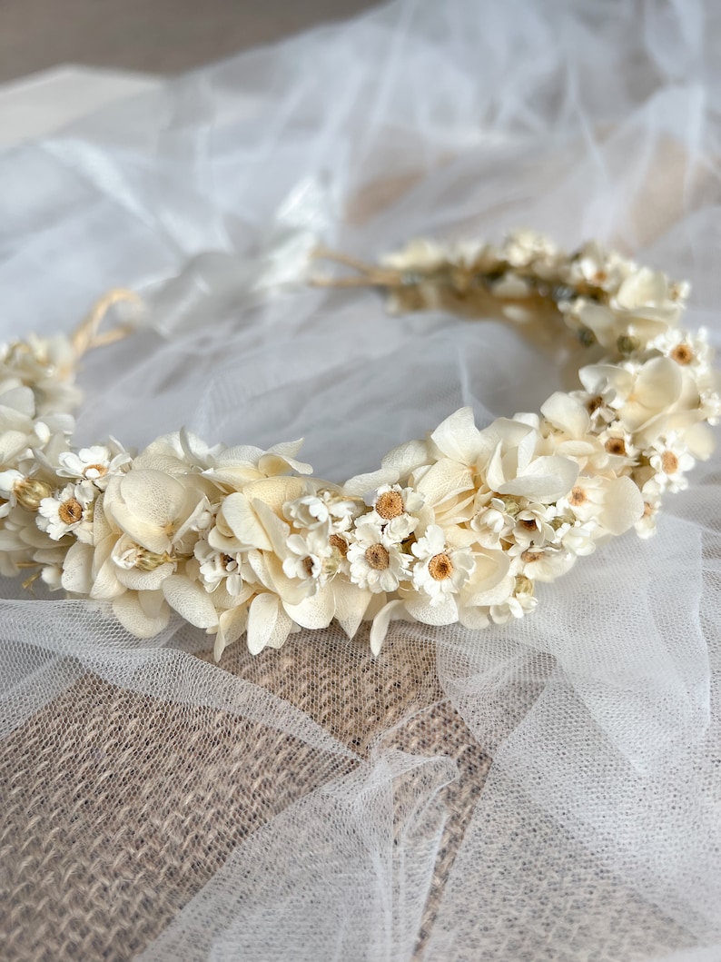 May include: A floral crown featuring an arrangement of cream and off-white dried flowers, including small daisies and hydrangea-like petals. The circular headpiece is constructed with a thin, flexible base and includes white ribbon ties at the back.