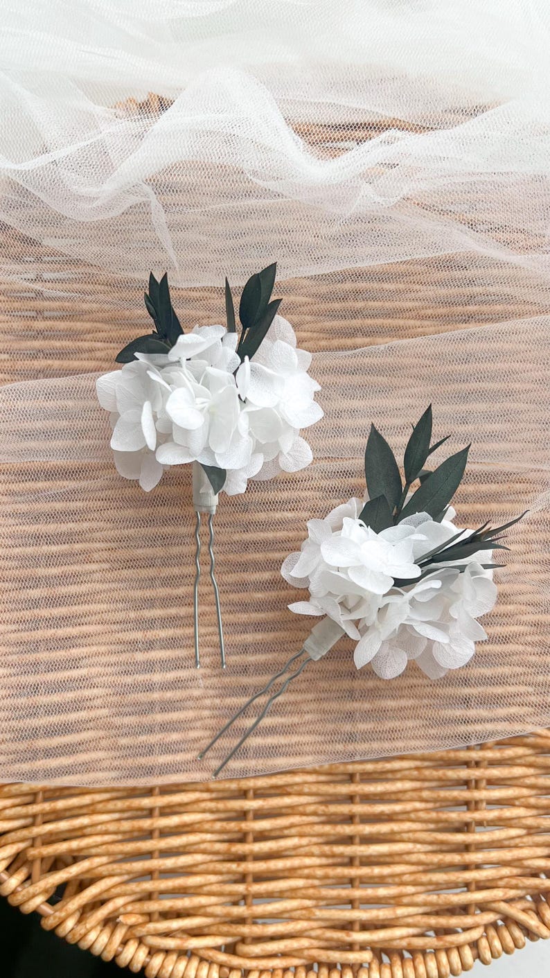 Puede incluir: Dos horquillas para el pelo con racimos de p&eacute;talos de hortensia blancos acentuados con hojas de color verde oscuro. Cada arreglo floral est&aacute; fijado a una horquilla de metal plateado en forma de U, dise&ntilde;ada para colocarla en el cabello.